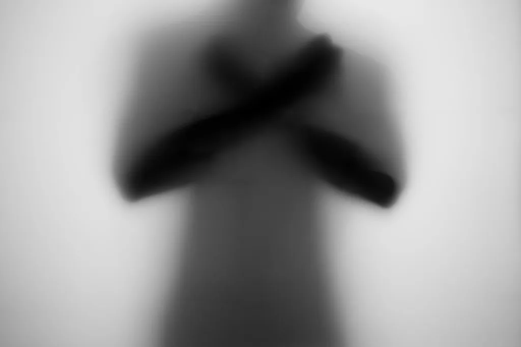 A blurred silhouette standing with both arms crossed over the chest, seen through frosted glass.