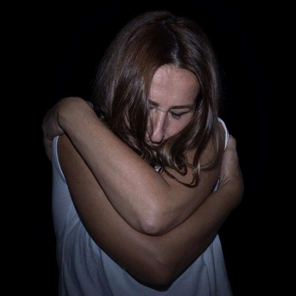 A woman in a white top hugs herself with both arms, head bowed and hair falling over her face, against black.