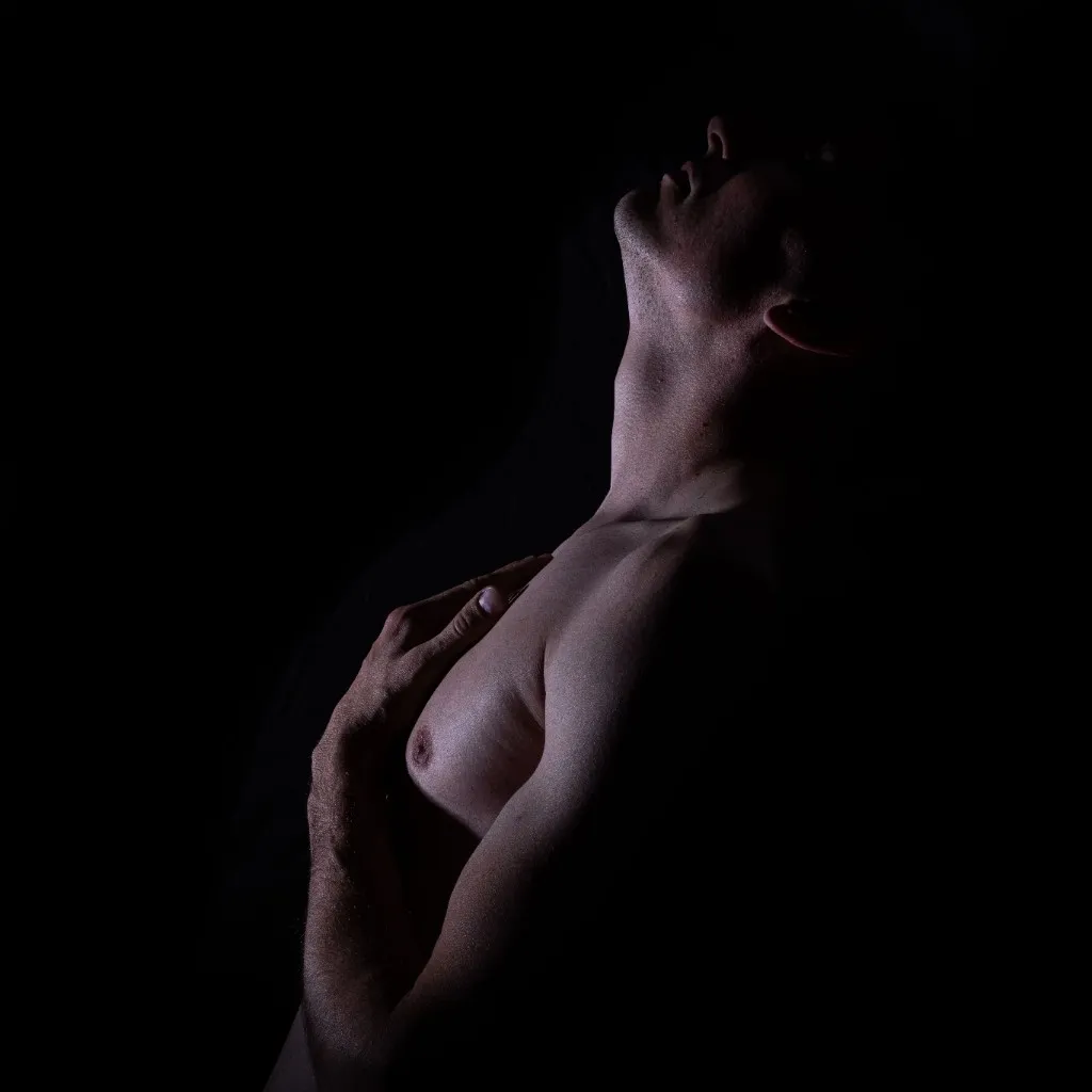 A bare torso arches back into darkness, head tilted up, one hand held over the heart.
