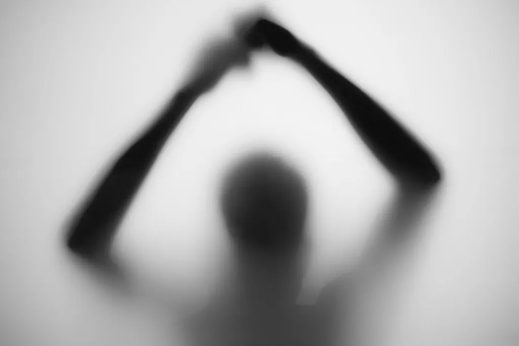 A silhouette with both arms raised overhead, elbows out, forming an arch above a bowed head.
