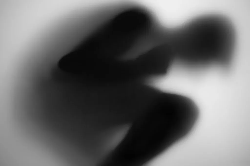 A body curled inward with knees drawn up, reduced to a soft dark mass behind glass.