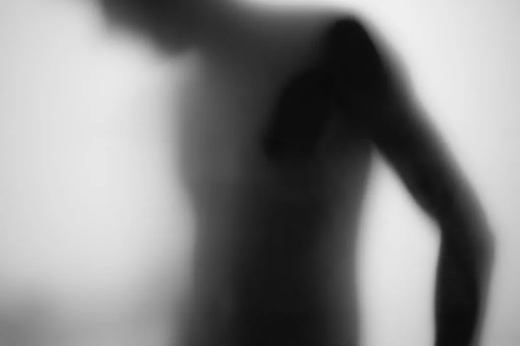 A bare torso turning in profile, one arm resting along the side, seen through frosted glass.