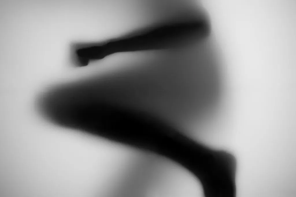Folded legs pressed to frosted glass, knees and shins blurred into soft dark curves.